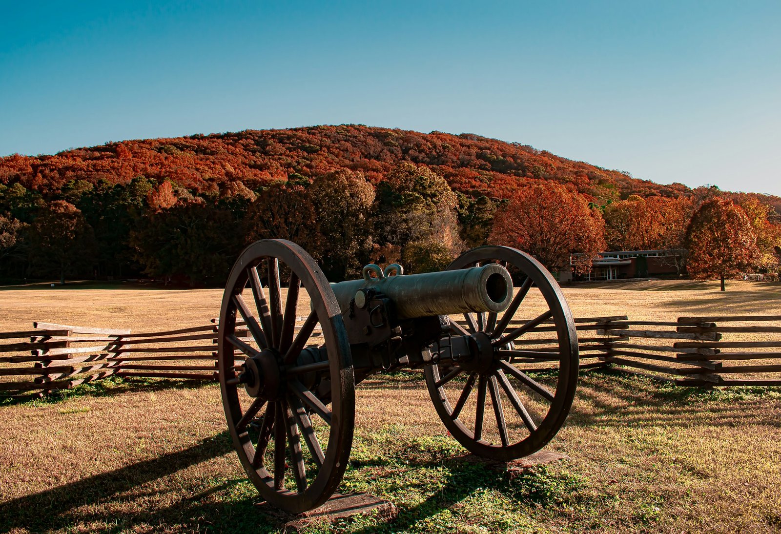 A Civil War cannon behind a split-rail fence below Kennesaw Mountain in autumn, with the park Visitor Center at right