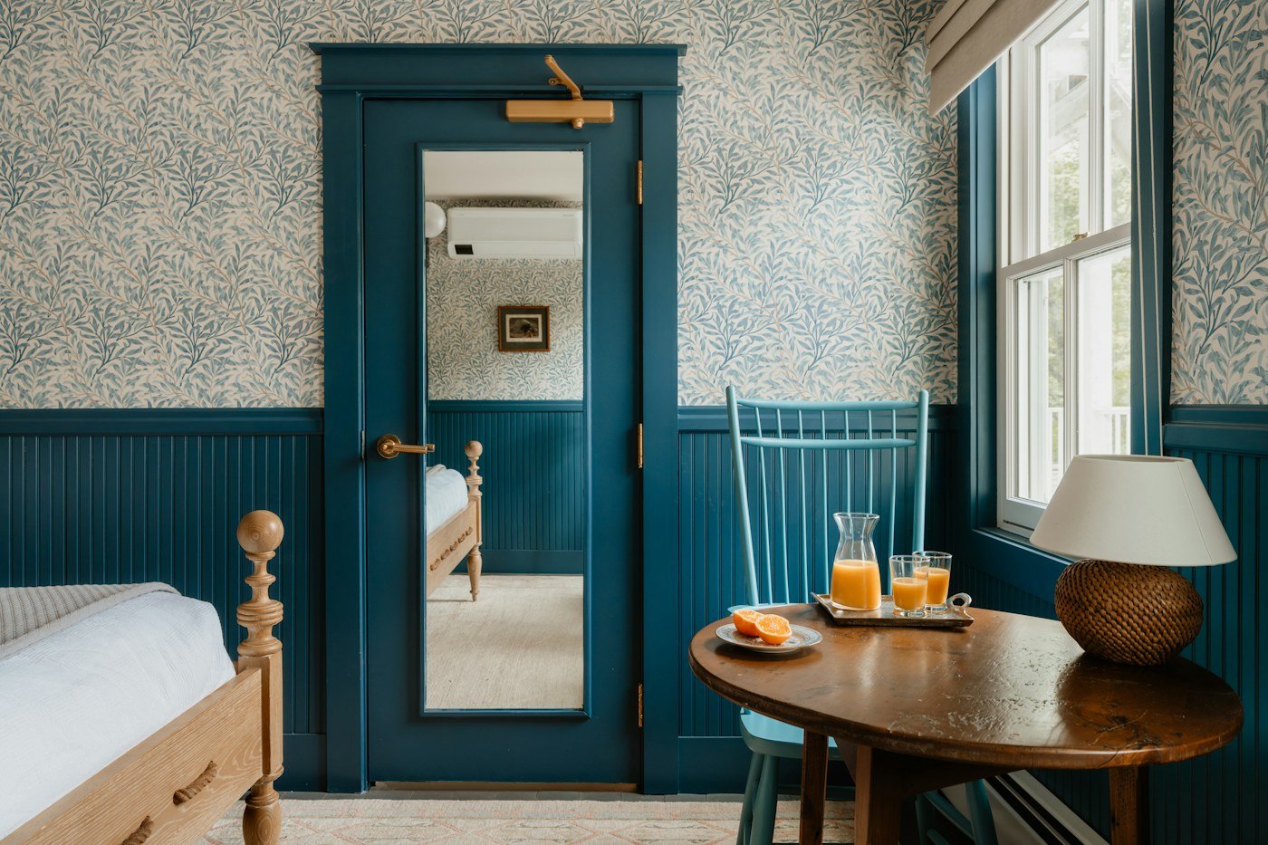 A small historic-style hotel room with leaf wallpaper, blue wainscoting, a wooden bed and a table by the window