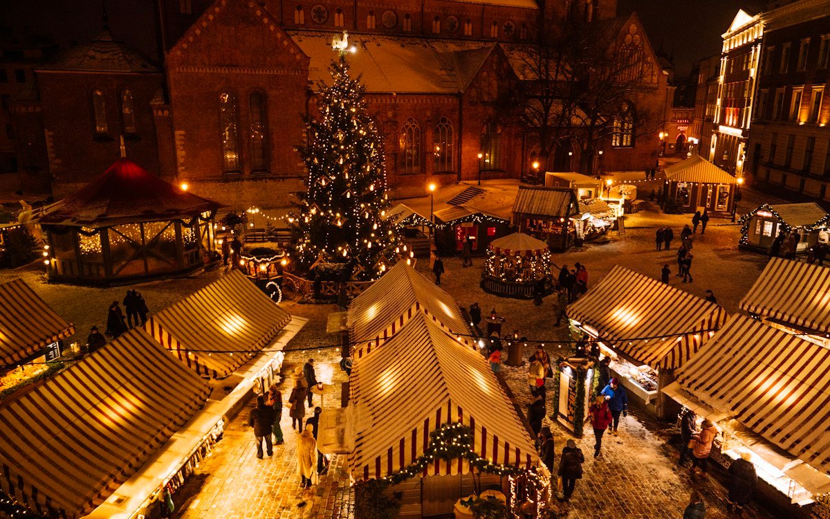 An evening view of a Christmas market with striped-roof stalls, a lit tree and a crowd