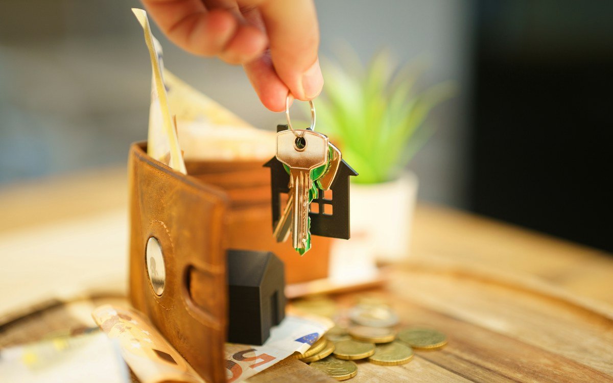 House keys with a small house charm resting on a leather wallet beside coins