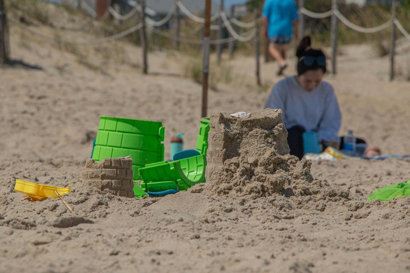 A sandcastle, green buckets and a yellow shovel on a sunny beach with a parent sitting out of focus behind (illustrative, not Tybee Island).