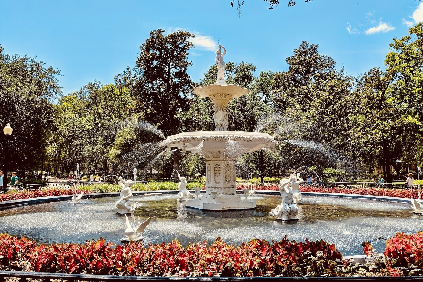 The white Forsyth Park fountain in Savannah, Georgia, spraying water above red flower beds under a blue sky and oak trees.