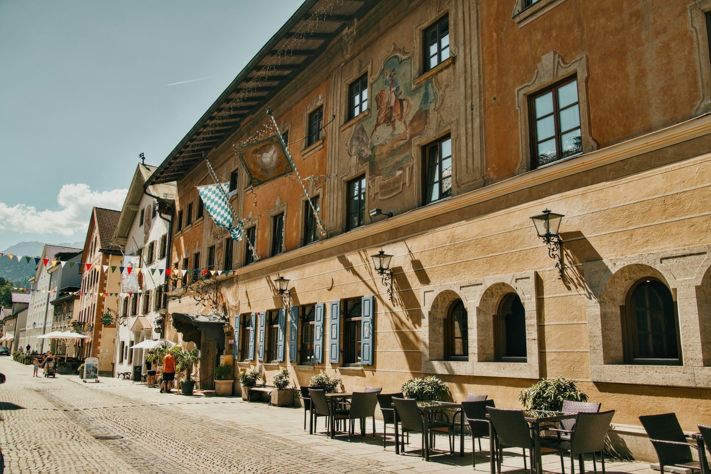 A cobblestone street lined with painted Alpine-style buildings, cafe tables and small flags.