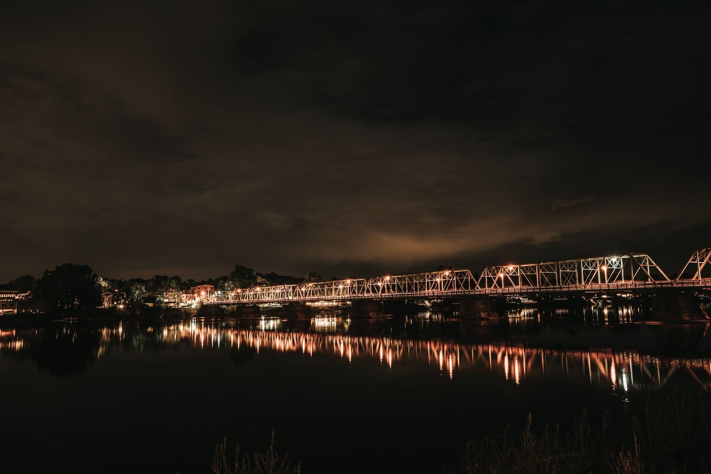 A lit steel truss bridge reflected in still river water at night under cloudy skies.