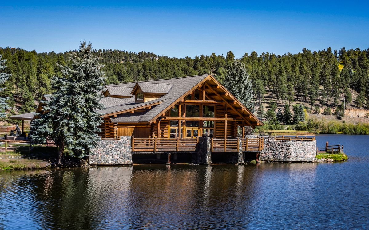 A large log lodge with a deck beside a pond among forested hills