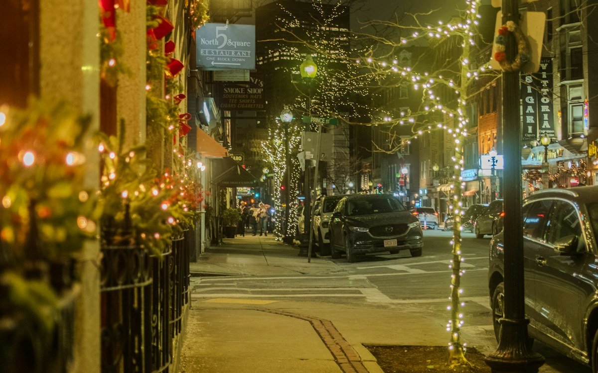 A city street at night with Christmas lights wrapped around trees and lamp posts