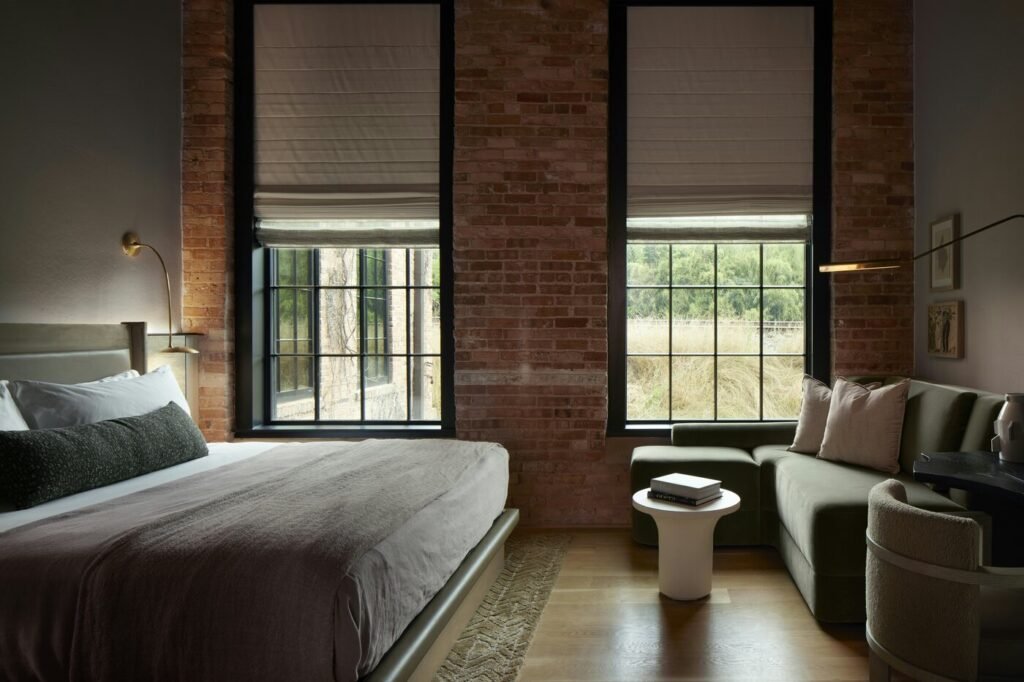 A Premier King room at Rivet House in Athens, with exposed brick walls, two tall black-framed windows, a king bed and a green sofa